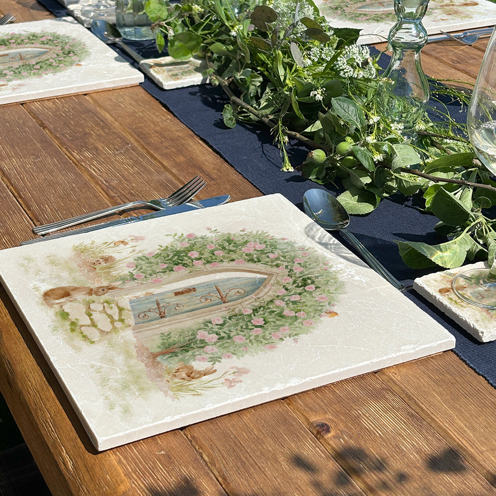 A handmade marble placemat with a magical watercolour scene of a hidden garden door under an archway of roses. The placemat is set on a beautiful garden dining table.