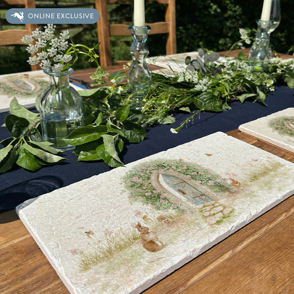 A rectangle marble sharing platter with a watercolour scene showing a hidden door under a rose archway leading to a secret garden. The platter is set on an al fresco table with matching placemats.
