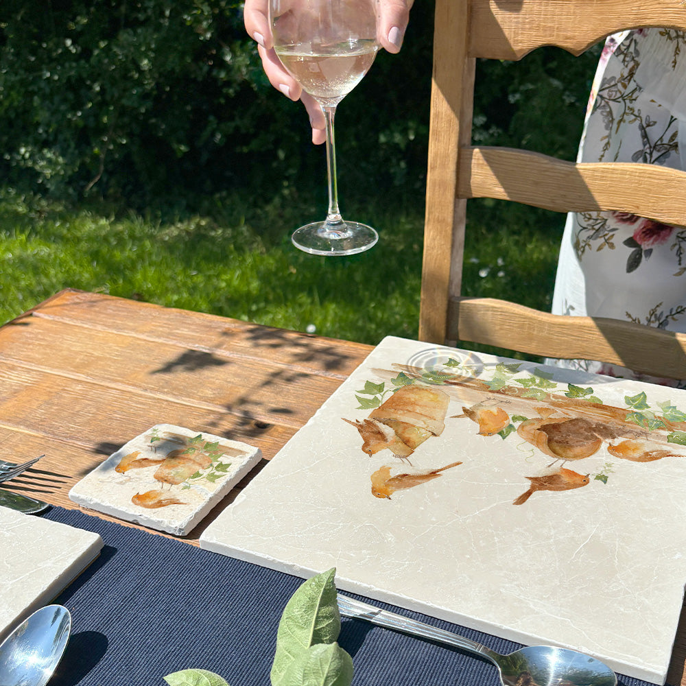 A sunny outdoor table set with marble placemats and coasters, a hand is about to place a glass of wine on the coaster which features a robin watercolour design.