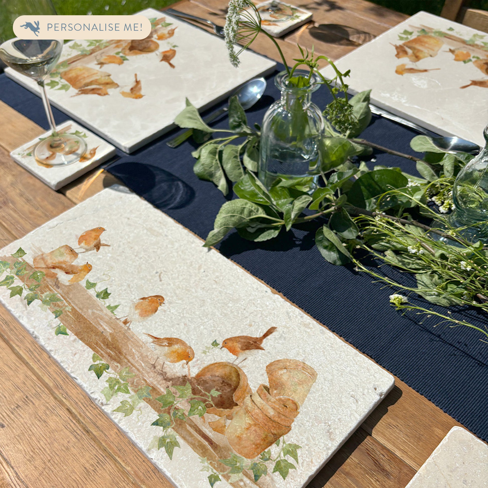 An outdoor garden table set with marble platters and coasters, each featuring a watercolour design of robins up to mischief on a shelf full of plant pots.