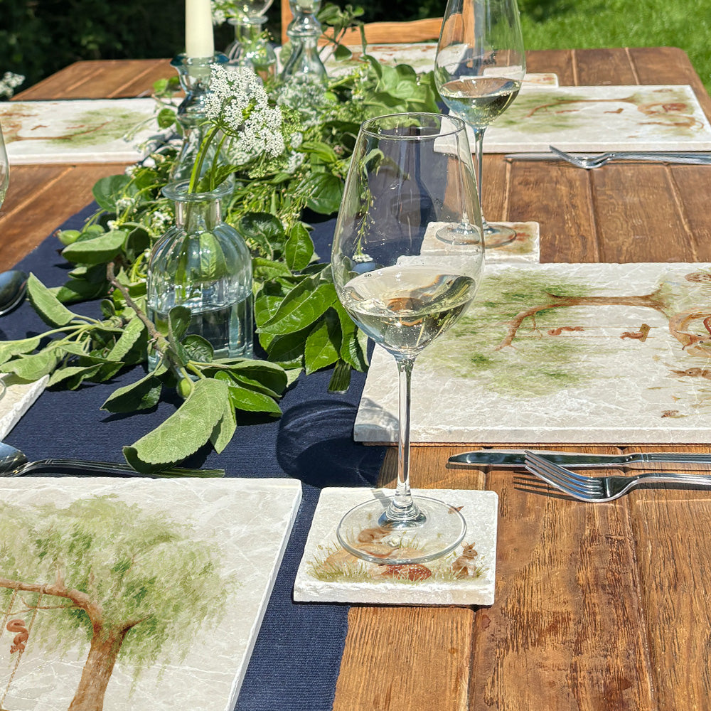 An outdoor dining table set with marble placemats and coasters featuring a watercolour scene of woodland animals under a willow tree.