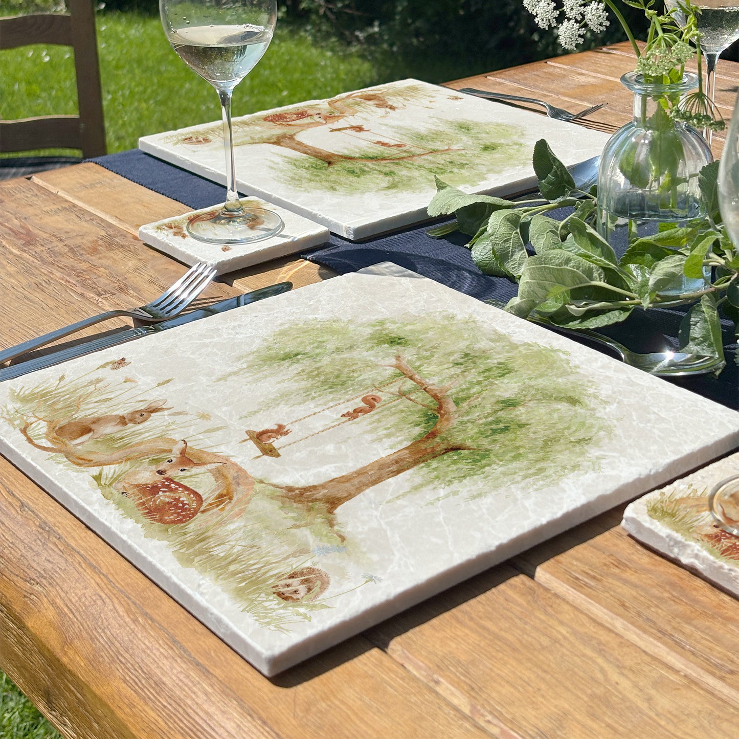 A garden table set with large marble platters featuring a watercolour scene of woodland animals under a willow tree.