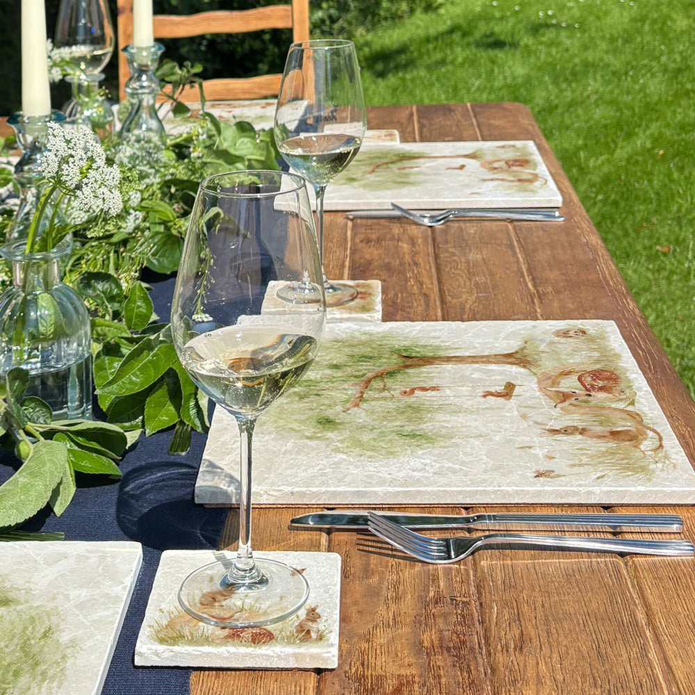 A garden table set with large marble placemats featuring a watercolour scene of woodland animals under a willow tree.