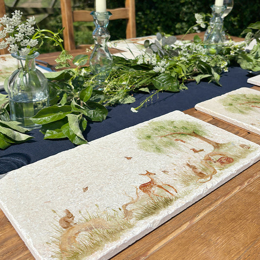 A handmade marble serving platter featuring a watercolour scene of woodland animals under a willow tree. The platter is set on an outdoor garden table.