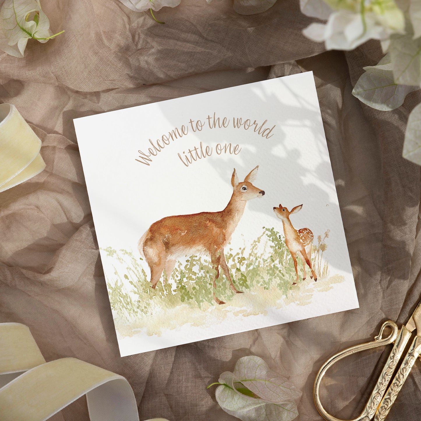 A greetings card laid flat on a table surrounded by gift wrapping items including scissors and ribbon. The card reads Welcome to the World Little One in brown text above a red deer doe and baby fawn in a watercolour style.
