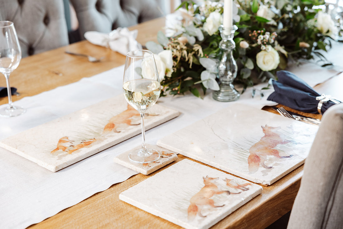 A wooden farmhouse style table set with marble placemats and coasters that feature a watercolour design of a fox and fox cub.