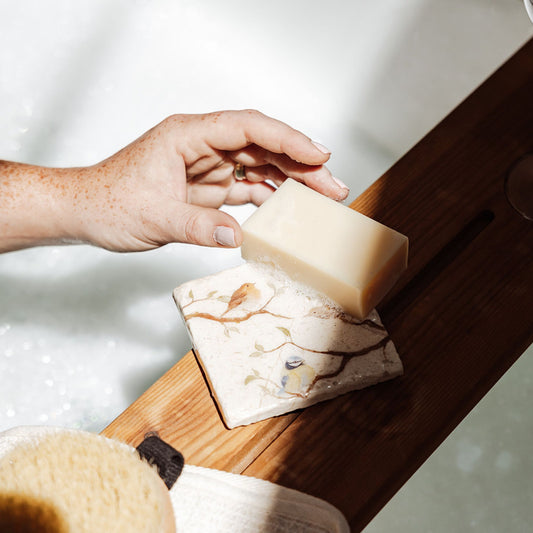 A square marble coaster being used as a soap dish on a wooden bath tidy above a bubble bath. The coaster features a British garden birds watercolour design.