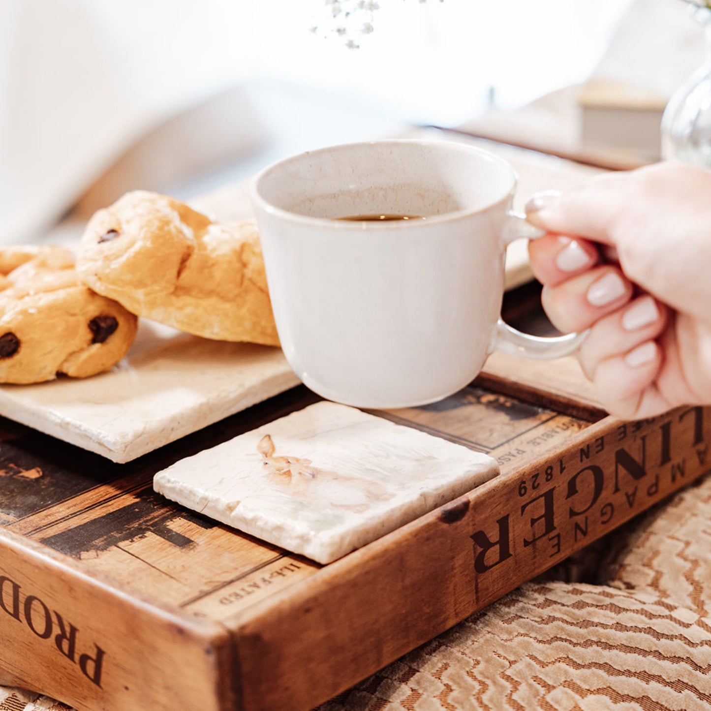 A hand holding a cup of coffee, about to place it onto a square marble coaster on a breakfast tray. The coaster has a watercolour design featuring a hare washing his ear.