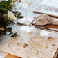 A wooden farmhouse dining table set with large square marble placemats. The placemats feature bees and a beehive in a buttercup meadow, in a watercolour style.