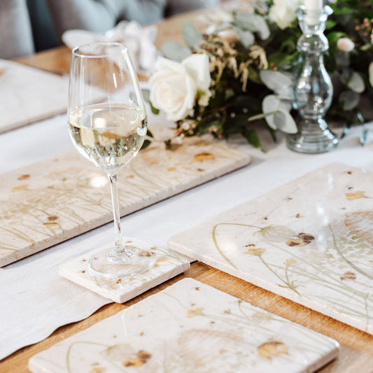A wooden dining table set with cream marble placemats and coasters. The placemats are square and feature bees and a beehive in a buttercup meadow, in a watercolour style.