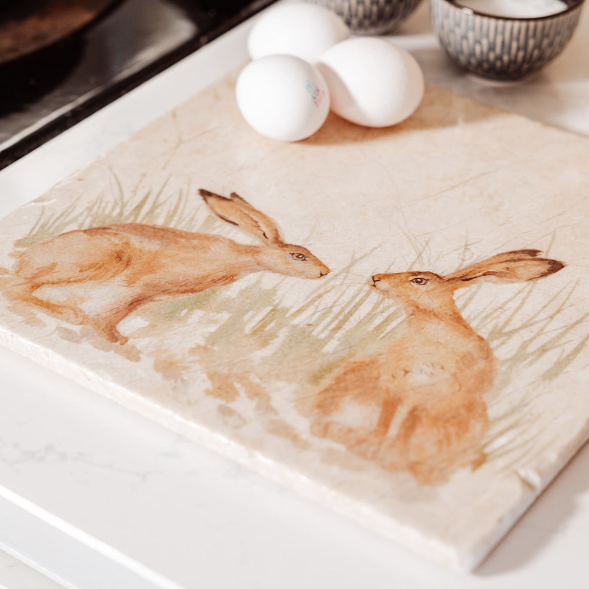 A large square marble platter on a country kitchen worksurface. The platter features a watercolour design of two hares facing each other about to touch noses.