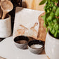 A medium square cream marble platter propped up on a country kitchen worksurface among kitchen utensils and wooden chopping boards. The marble platter features a watercolour design of two hares facing each other about to touch noses.