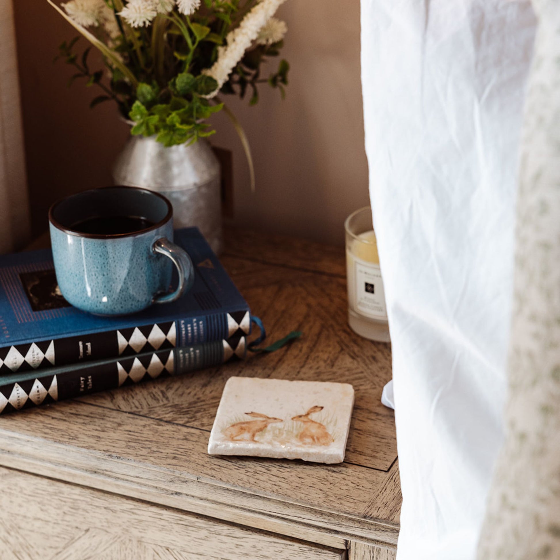 A square marble coaster on a bedside table next to a cup of coffee and a pile of books. The coaster has a watercolour design featuring two hares facing each other about to touch noses.