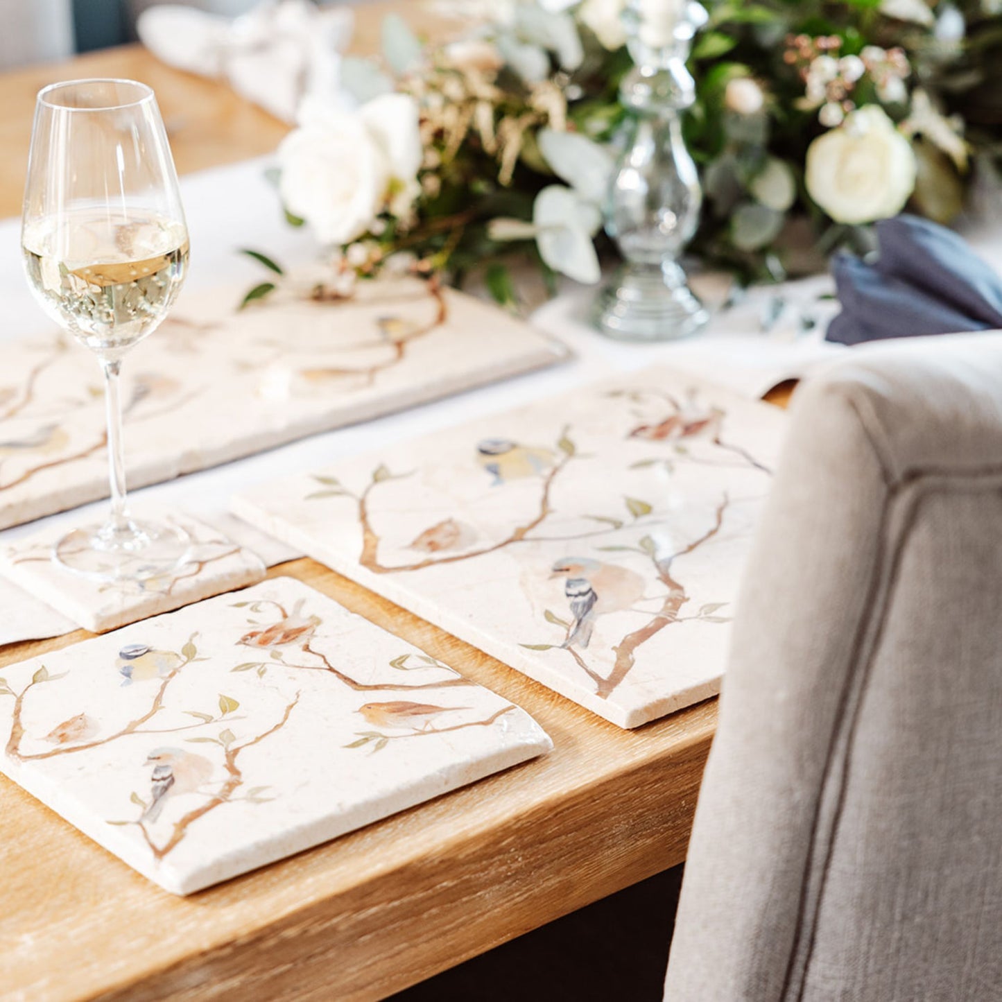 A wooden dining table set with cream marble placemats and coasters. The placemats are square and feature British garden birds on branches in a watercolour style.
