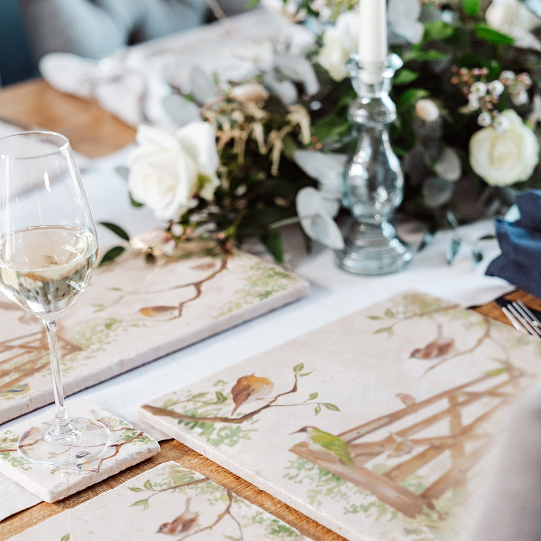 A wooden dining table set with cream marble placemats and coasters. The placemats are square and feature British garden birds on paddock gate in a watercolour style.