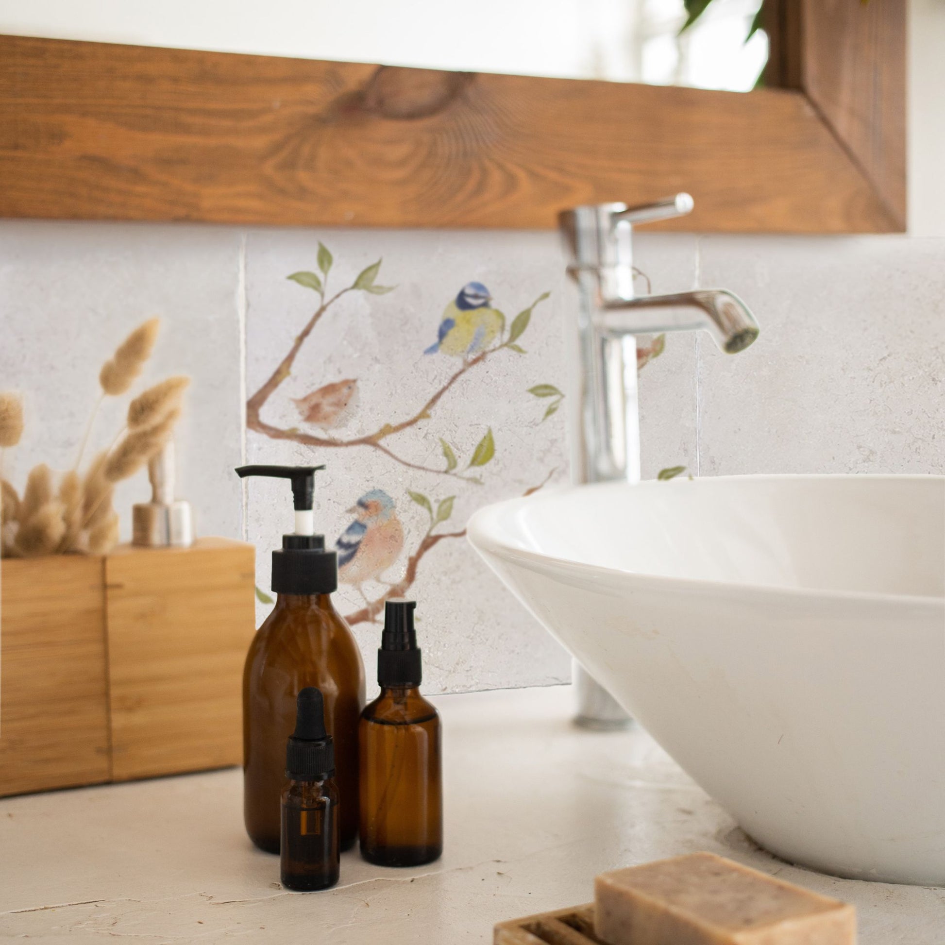 A tiled splashback behind a bathroom sink. The splashback is made up of plain cream square marble tiles alternated with tiles featuring countryside scenes including British garden birds on branches.