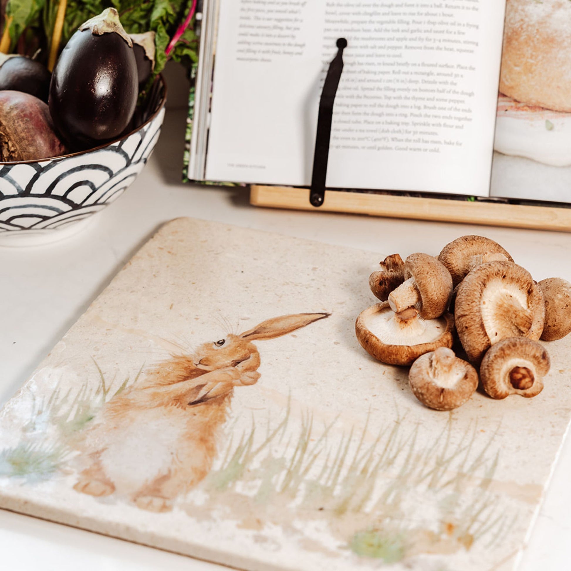A large square marble platter on a kitchen worktop with a recipe book propped up behind it. The platter has a hare design on it, and is being used as a worktop saver.