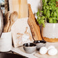 A medium square cream marble platter propped up on a country kitchen worksurface among kitchen utensils and wooden chopping boards. The marble platter features a watercolour design of two hares facing each other about to touch noses.