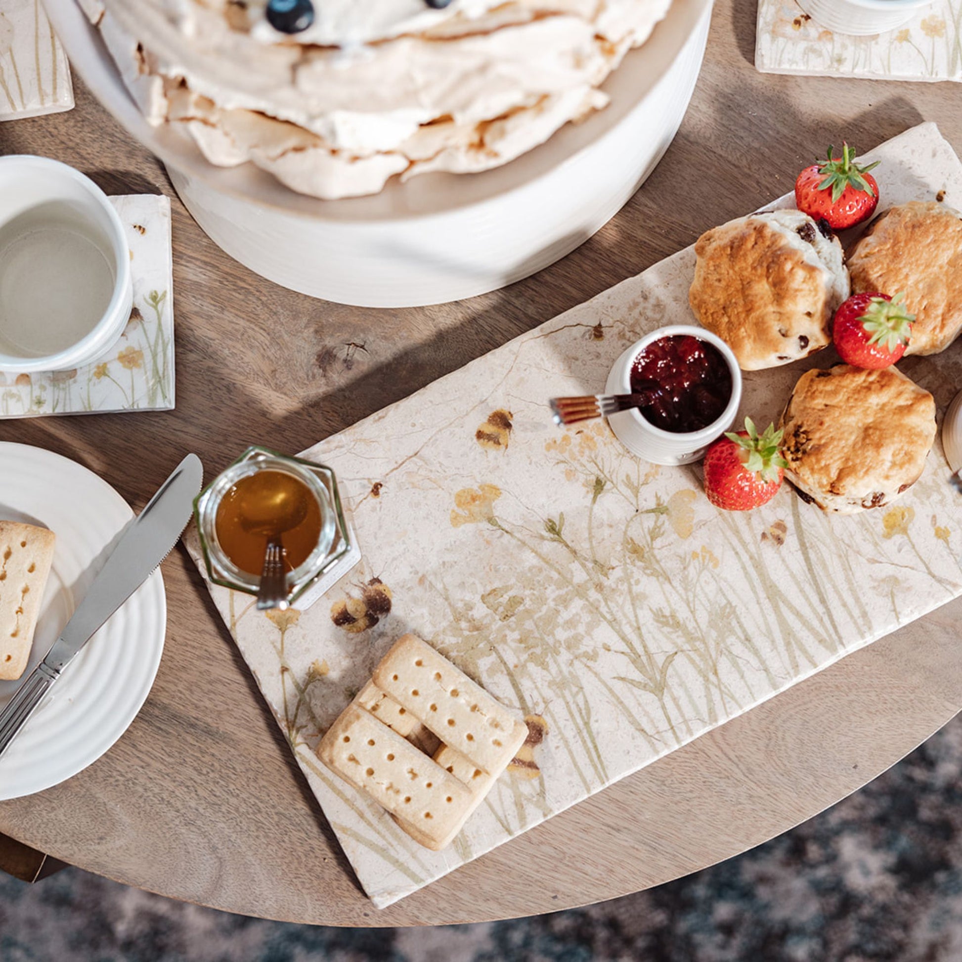 A rectangle marble sharing platter with a watercolour design featuring bees and a beehive in a buttercup meadow. The platter is being used to serve afternoon tea, including scones and jam.