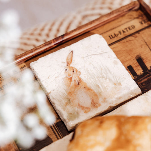 A square marble coaster on a breakfast tray. The coaster has a watercolour design featuring a hare washing his ear.
