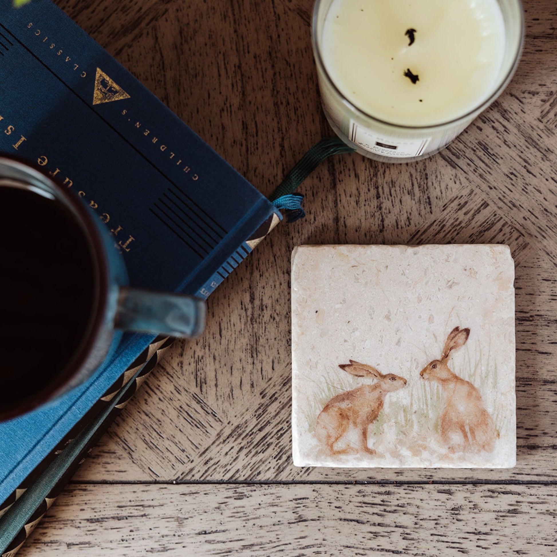 A square marble coaster featuring a watercolour design of two hares facing each other about to touch noses. The coaster is placed on a wooden table with a book, candle and cup of coffee.