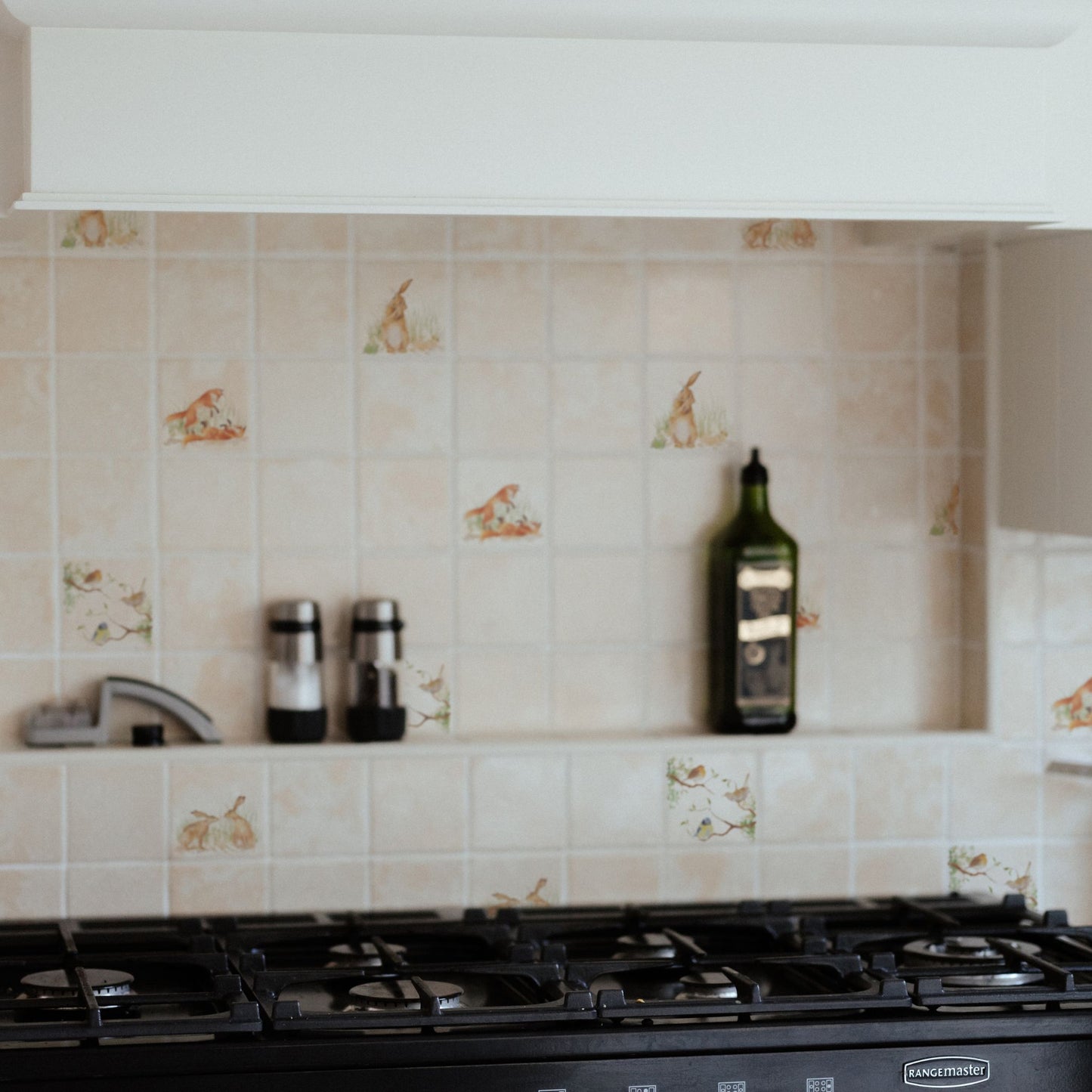 A tiled splashback behind a rangemaster oven in a country kitchen. It is made up of plain marble tiles, alternated with marble tiles featuring countryside animals including hares, foxes and garden birds. 