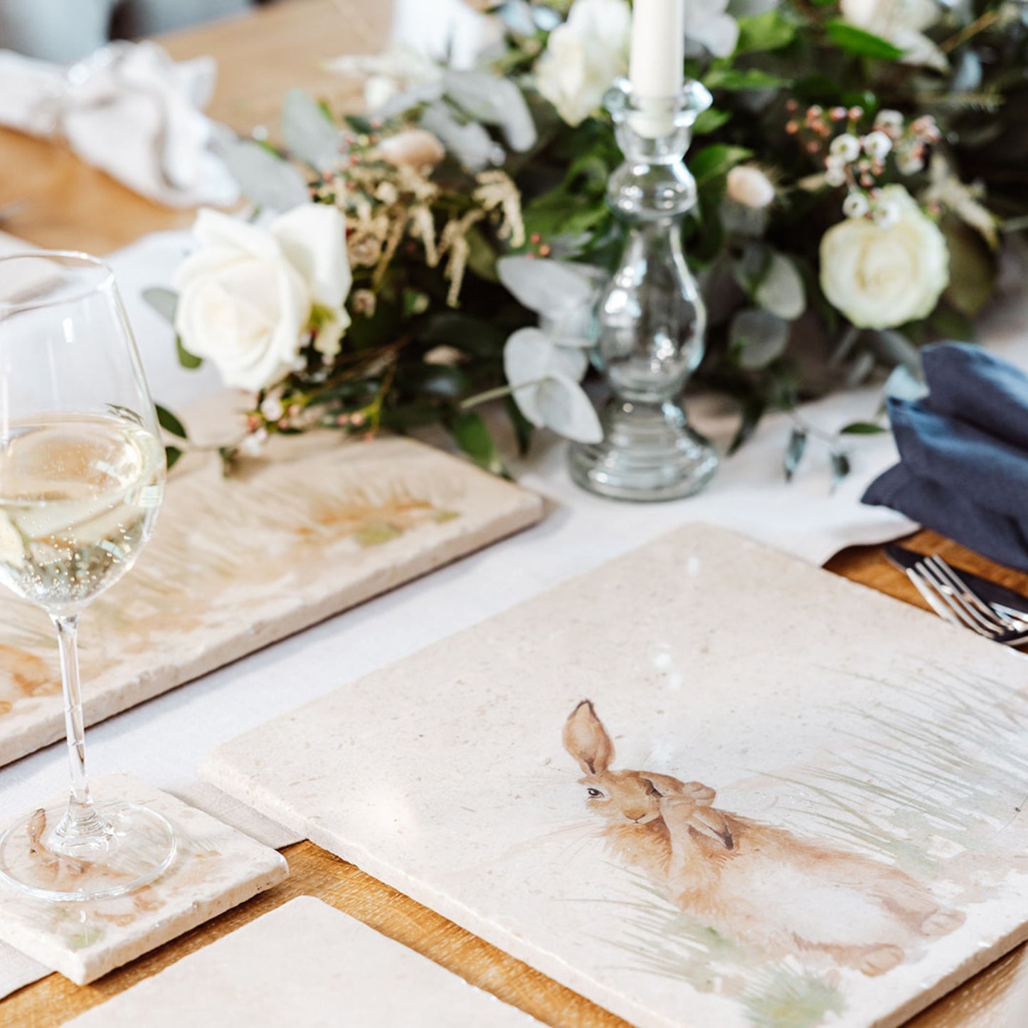 A wooden dining table set with cream marble placemats and coasters. The placemats are square and feature a hare in grass washing his ear in a watercolour style.