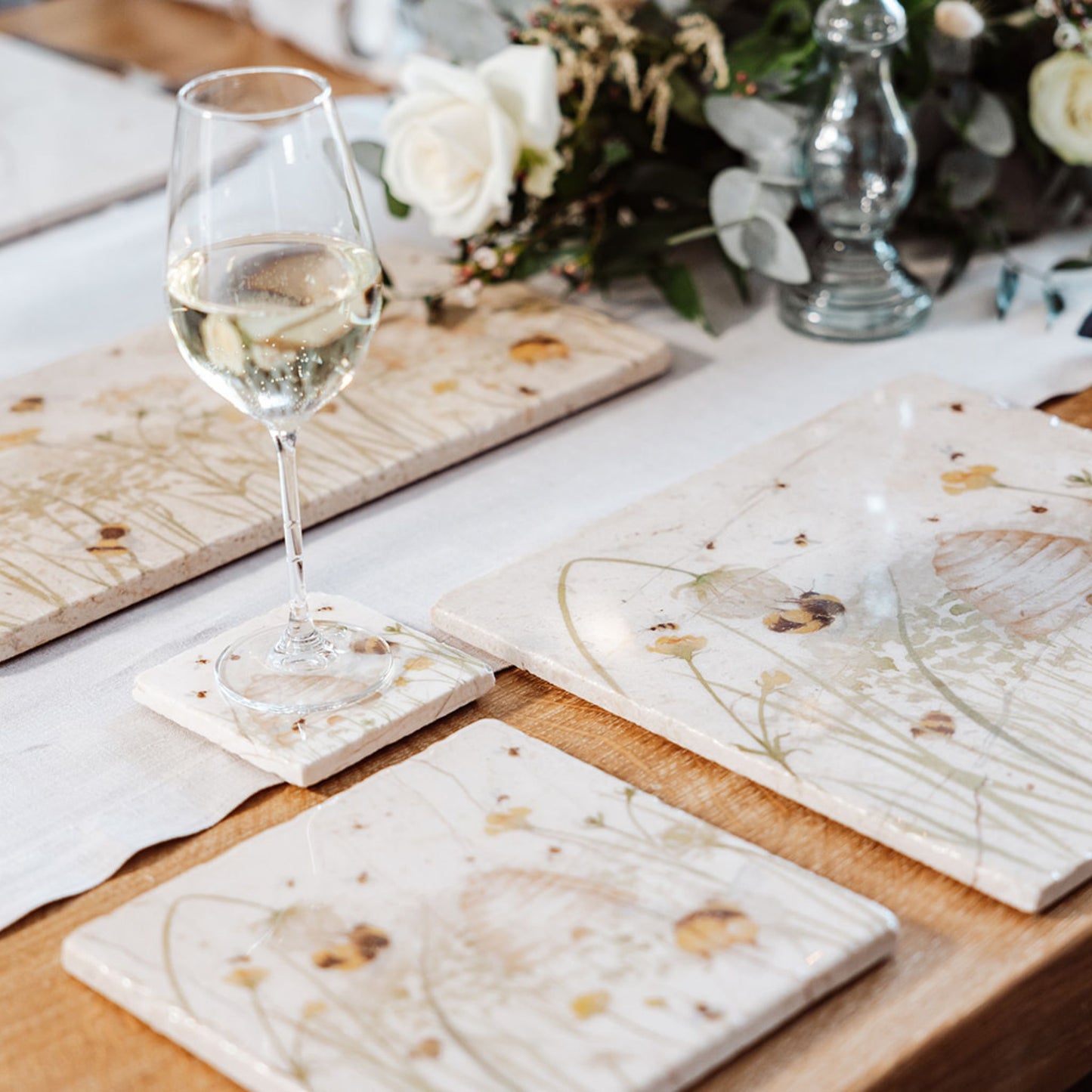 A wooden farmhouse dining table set with large square marble placemats. The placemats feature bees and a beehive in a buttercup meadow, in a watercolour style.
