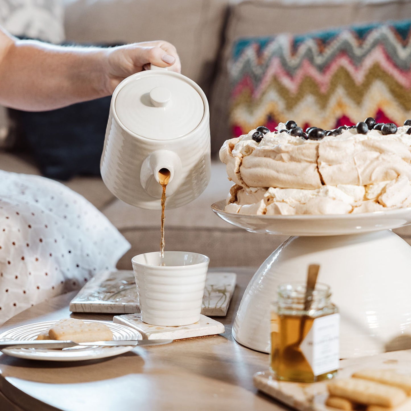 A table set for afternoon tea with marble platters and coasters. Tea is being poured from a cream teapot into a teacup resting on a marble coaster with a watercolour design featuring bees and a beehive.