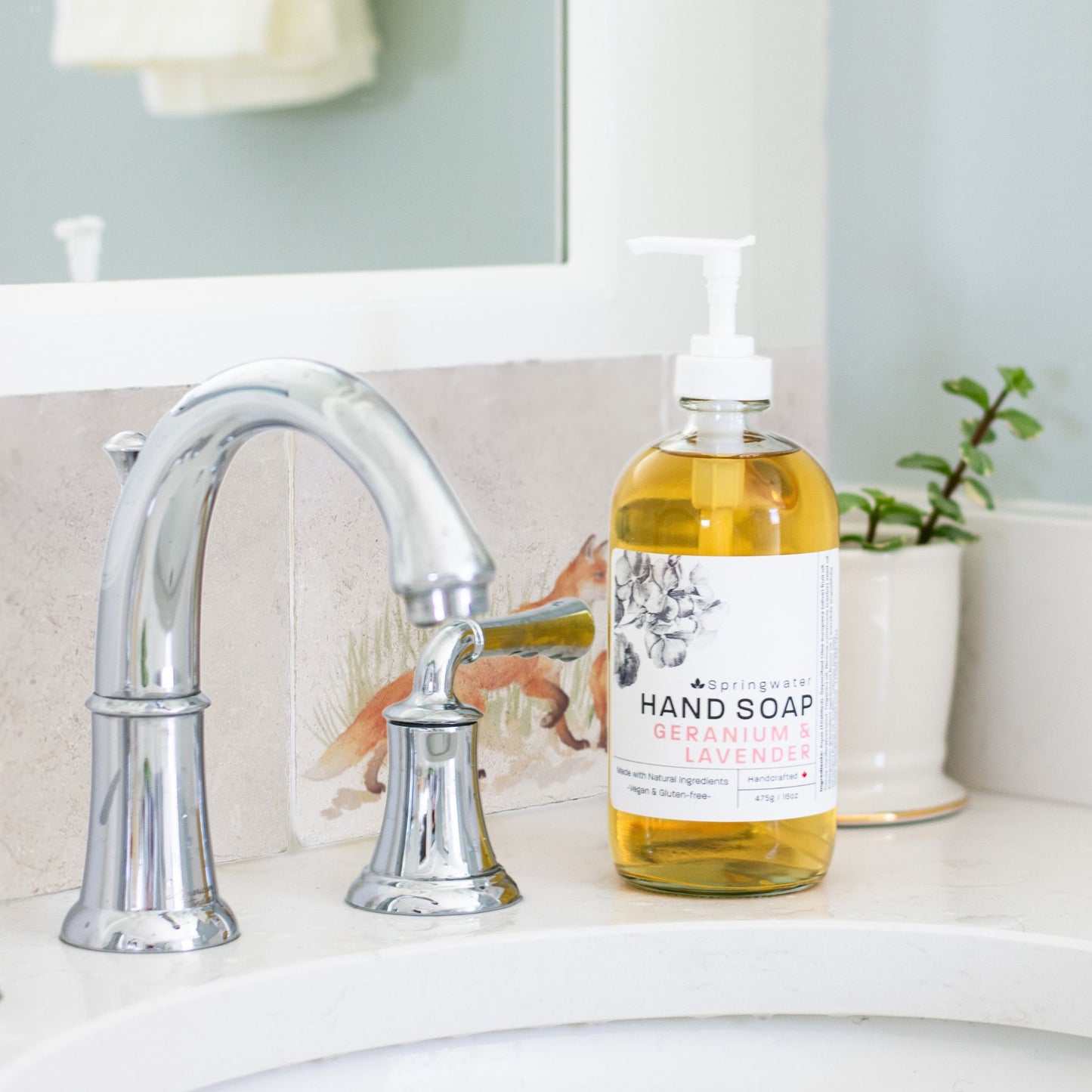 A tiled splashback behind a bathroom sink. The splashback is made up of plain cream marble tiles alternated with tiles featuring countryside scenes including a fox and fox cub.