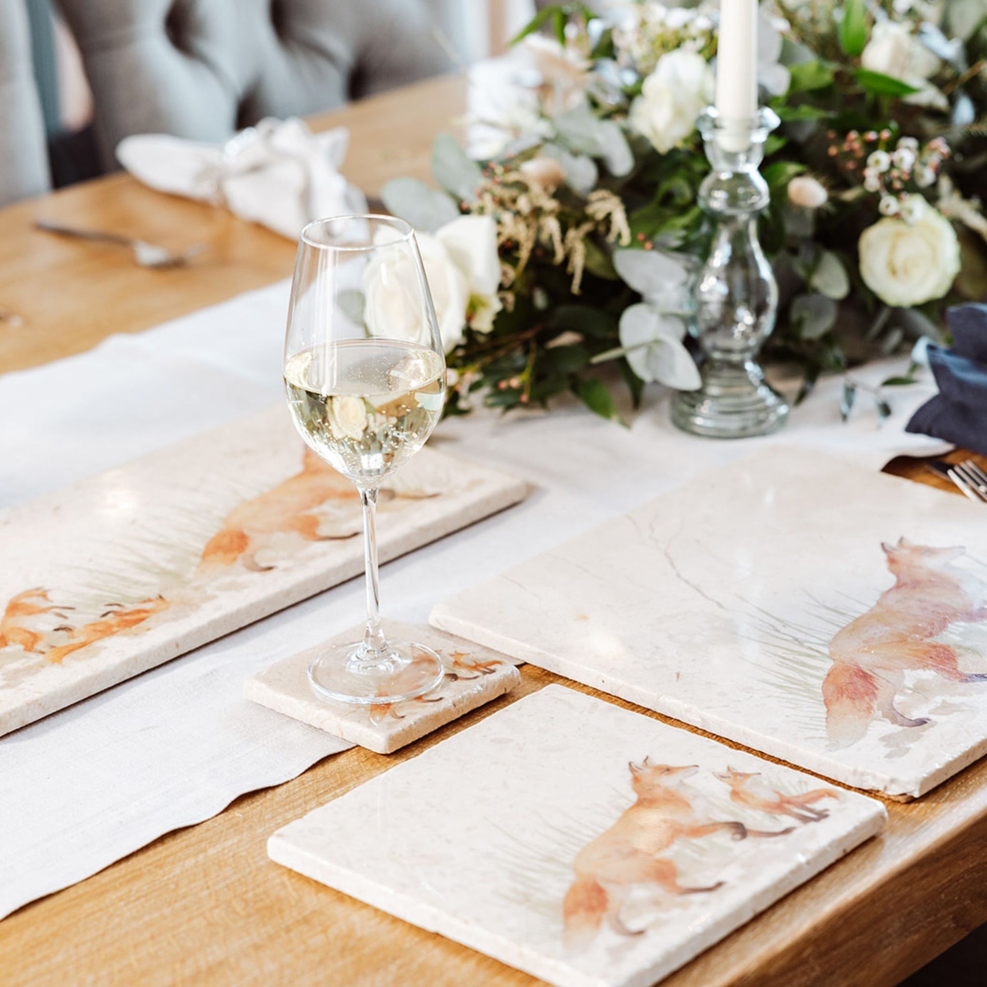 A wooden dining table set with cream marble placemats and coasters. The placemats are square and feature a fox and fox cubs in a watercolour style.
