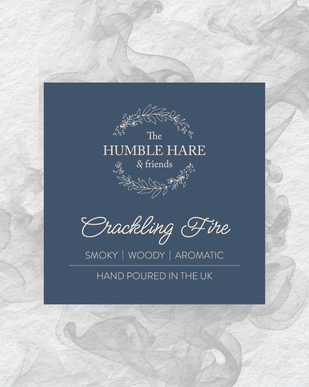 A square product label for 'The Humble Hare & friends' Crackling Fire' scented candle, featuring a dark blue background with elegant cream typography. The label is styled on a white textured background with a grey smoke effect behind it.