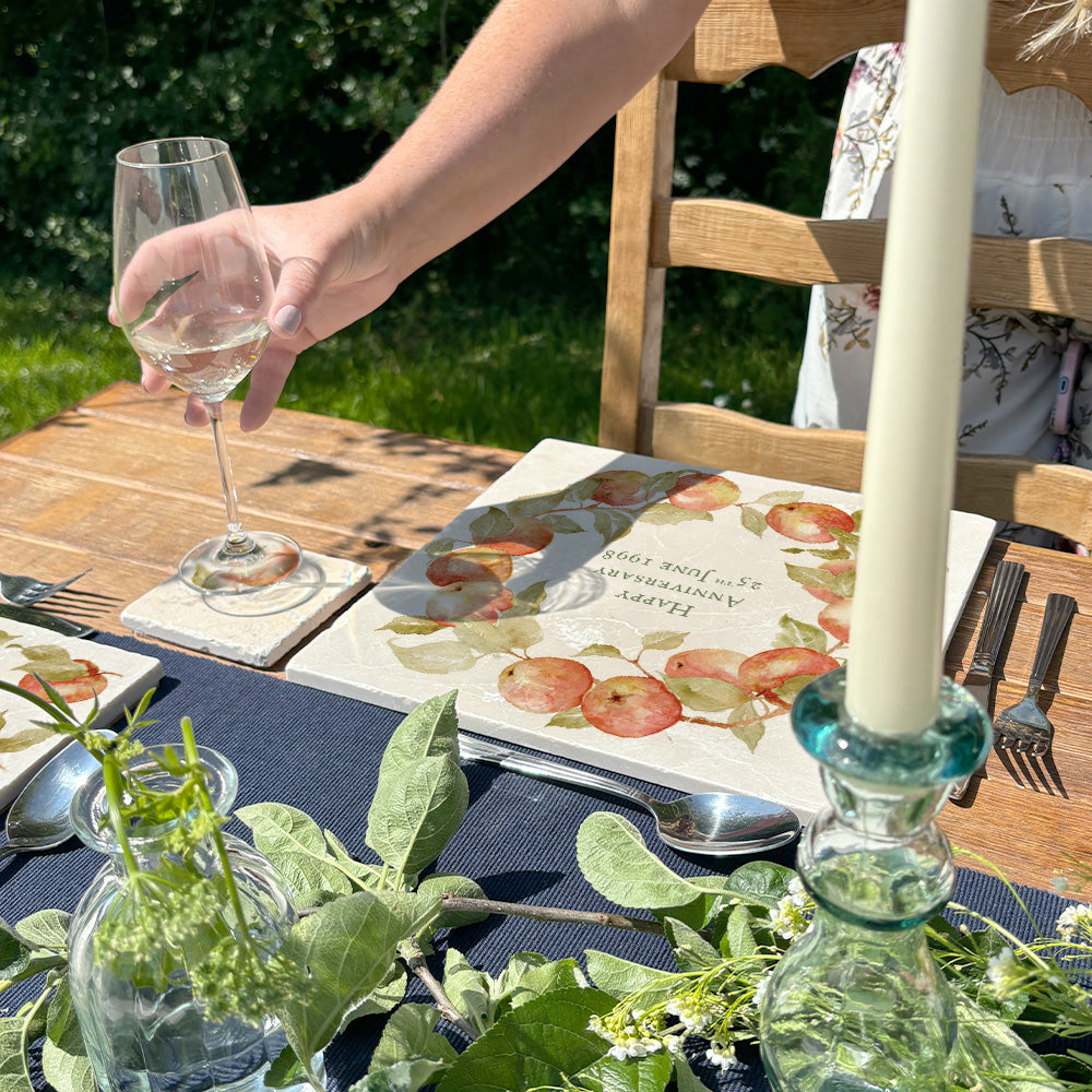 A personalised marble placemat with an apple and leaves wreath design, with a custom personalised message in the centre. The placemat is on a garden table set for a celebration dinner.