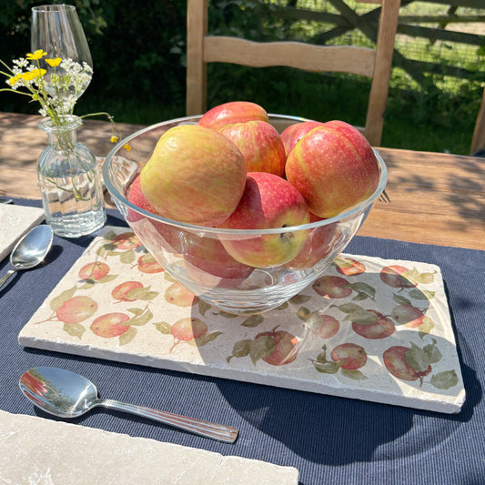 A large glass bowl full of ripe apples set as a dining table centrepiece on a rectangular marble sharing platter featuring a watercolour pattern of red apples and leaves.