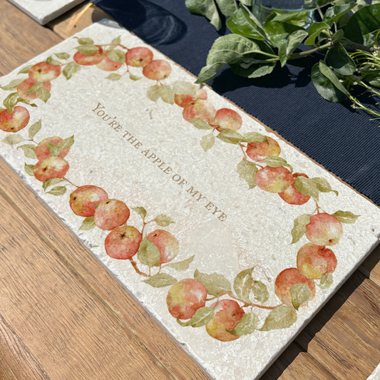 A personalised marble sharing board featuring a wreath of apples and leaves, with a bespoke message in the centre of the design. The custom sharing platter is set on an al fresco dining table decorated with foliage.