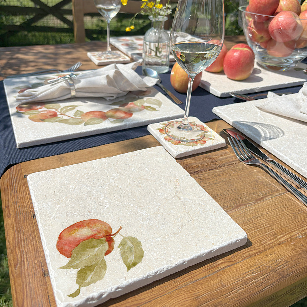 A garden table in the sunshine set with marble placemats, coasters and platters, featuring a watercolour design of a single apple with leaves.