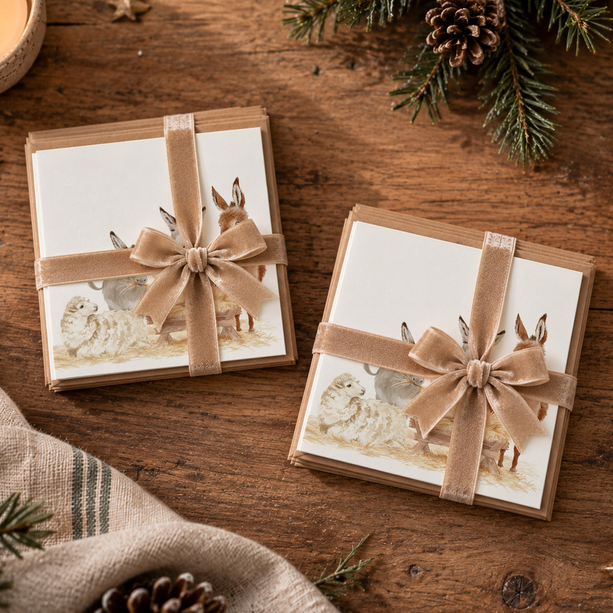 Two packs of Christmas cards wrapped with a champagne gold ribbon and bow, each printed with a rustic illustration of a nativity scene featuring sheep and donkeys, arranged on a wooden tabletop with pine branches and a candle nearby.
