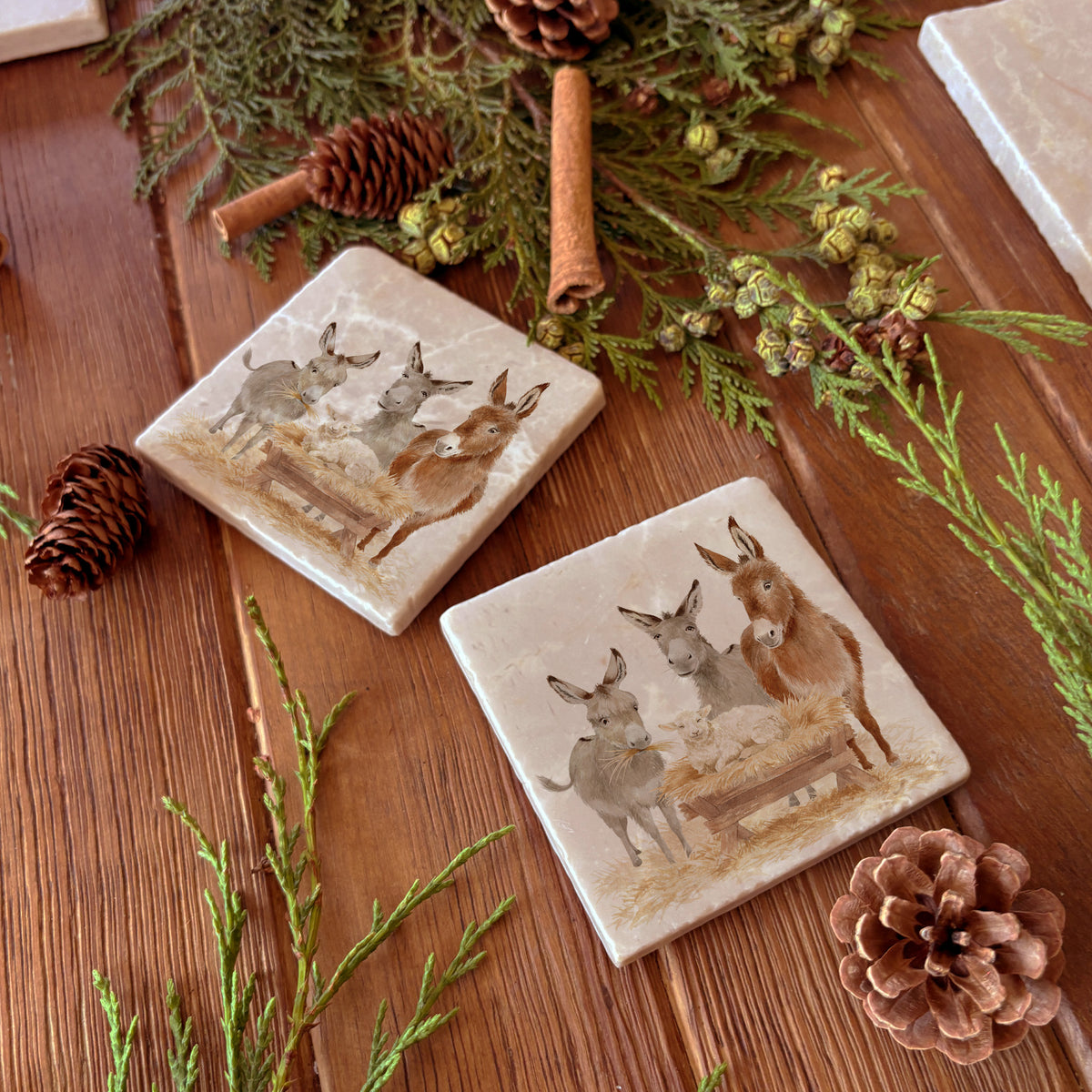 As set of four square marble coasters, each with a watercolour illustration of a nativity scene of donkeys and a lamb in a manger.