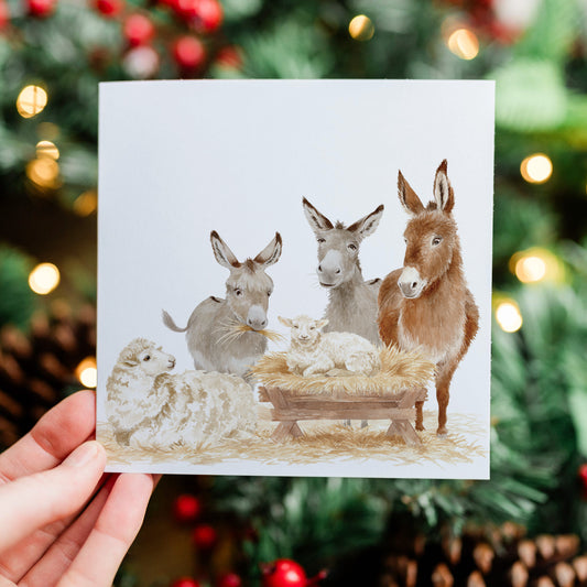 A hand holds a square greeting card featuring a watercolor nativity scene with donkeys, sheep, and a baby lamb in a stable. The background is a blurred Christmas tree with warm lights.
