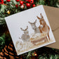 A square Christmas card featuring a watercolour nativity scene with donkeys, sheep, and a baby in a manger, placed on a brown envelope and surrounded by pine branches, red berries, and a pinecone.