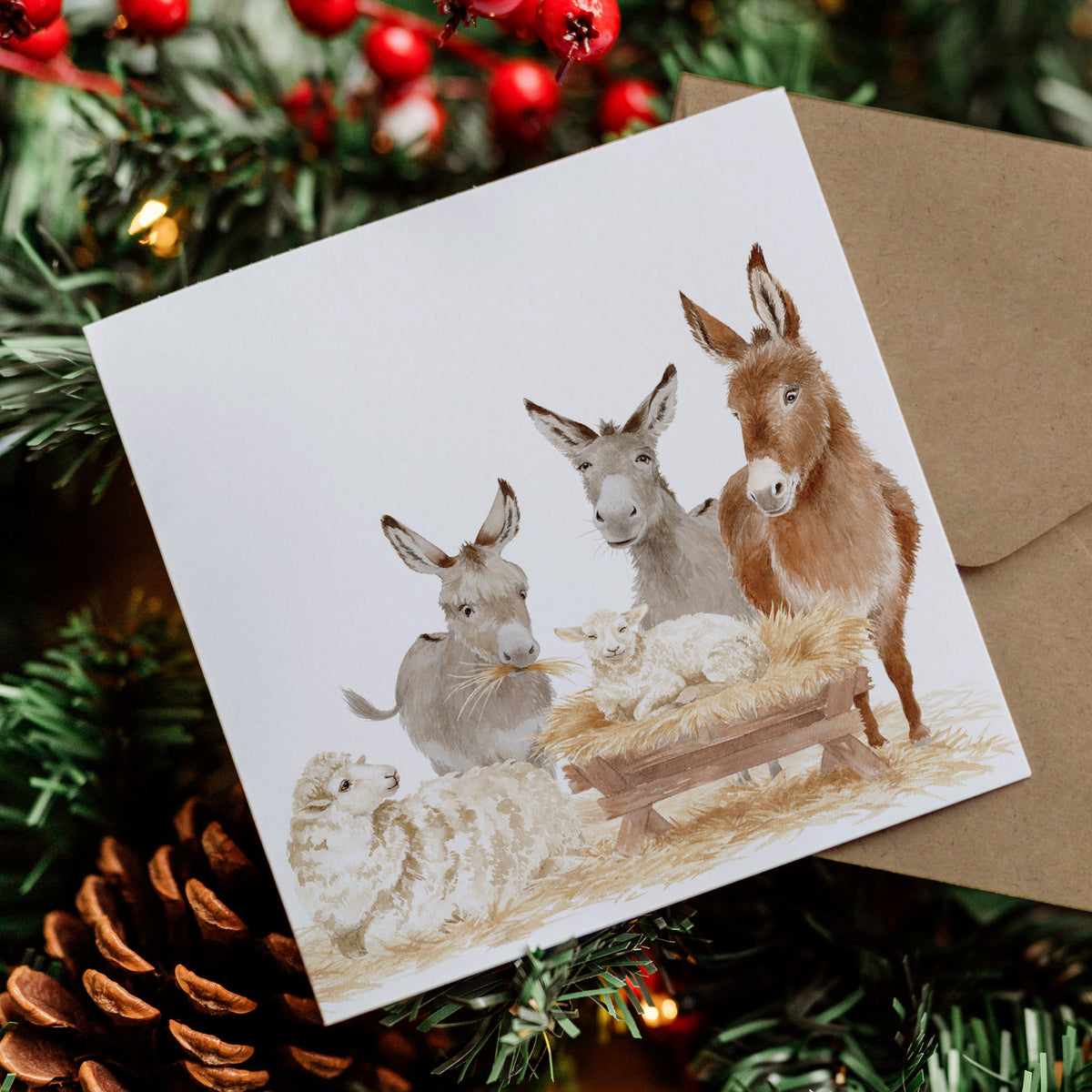 A square Christmas card featuring a watercolour nativity scene with donkeys, sheep, and a baby in a manger, placed on a brown envelope and surrounded by pine branches, red berries, and a pinecone.