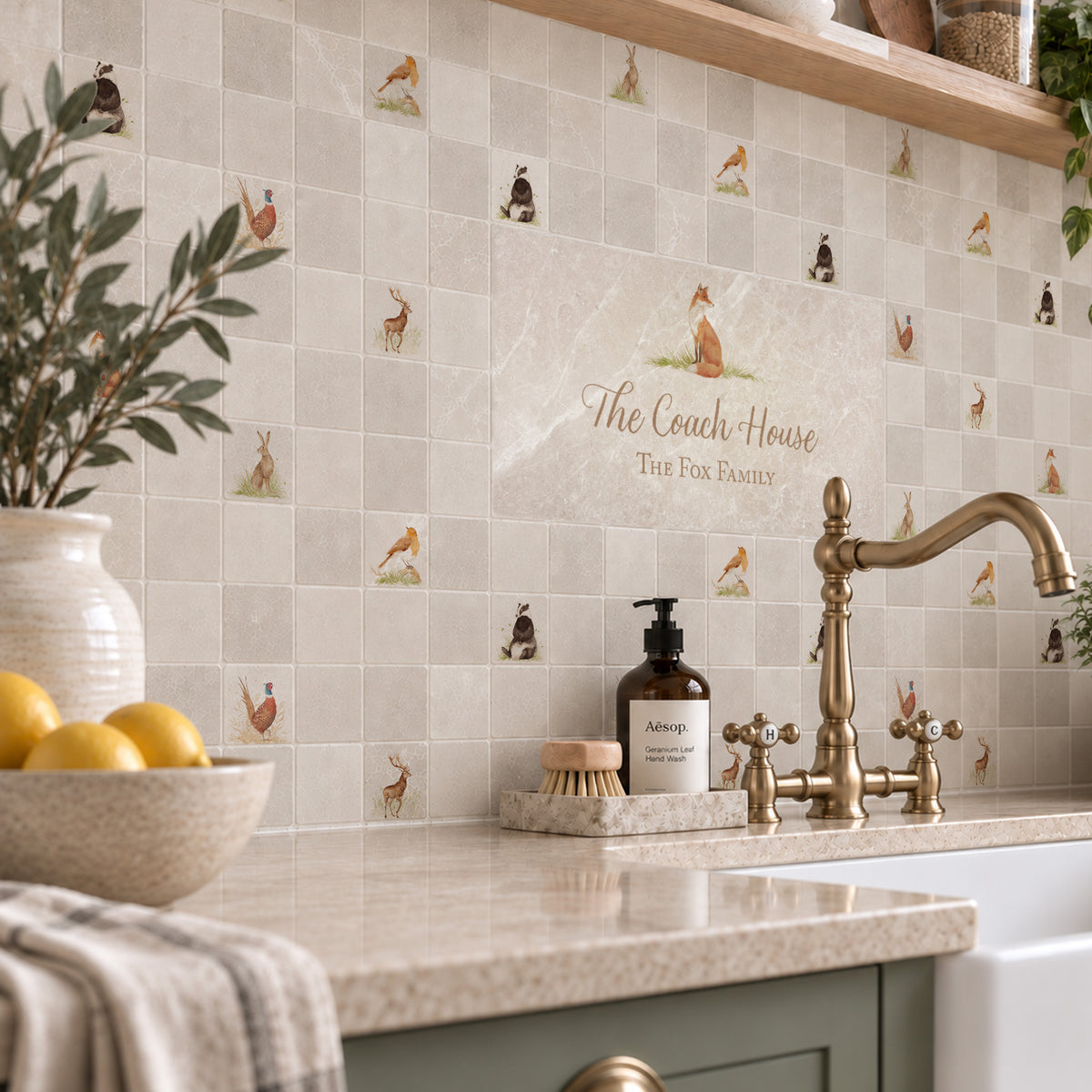 A kitchen backsplash made of square beige tiles with small illustrated foxes and rabbits, plus a central tile reading 'The Coach House The Fox Family.' A countertop with a brass faucet, soap dispenser, brush, bowl of lemons, and a vase is visible in the foreground.