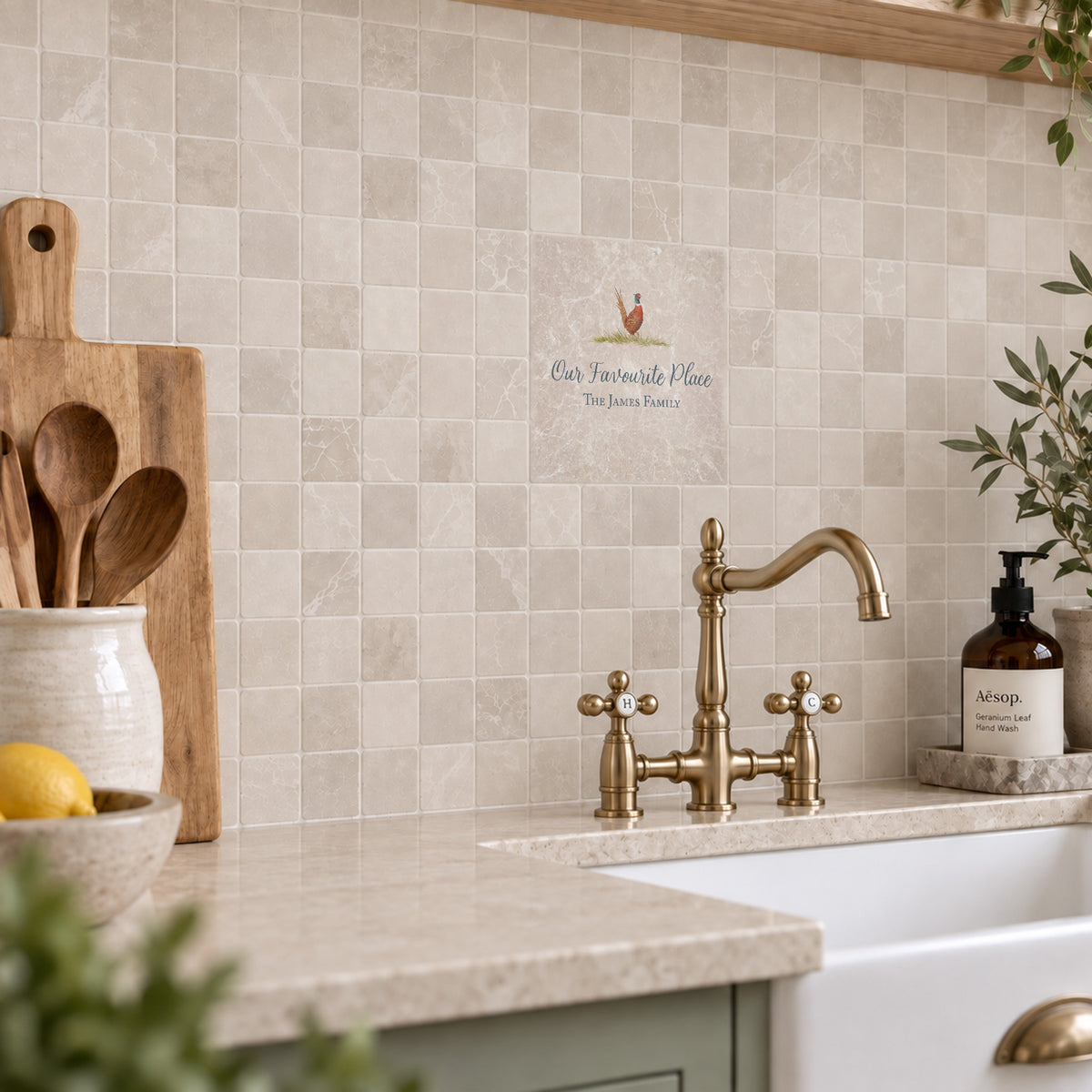 Beige marble-look square mosaic tiles on a kitchen backsplash behind a brass faucet and white farmhouse sink, with wooden utensils and a soap dispenser nearby.