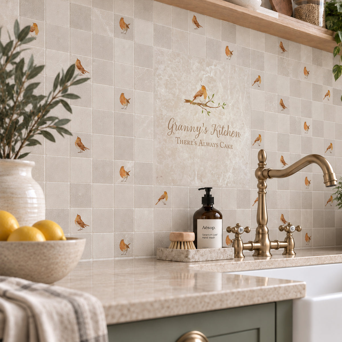 A styled kitchen interior featuring beige square mosaic tiles with a subtle repeating Robin pattern on the backsplash. The kitchen has sage green cabinets, a black induction cooktop, and wooden floating shelves with decorative ceramic vases and plants.