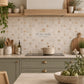A styled kitchen interior featuring beige square mosaic tiles with a subtle repeating Hare pattern on the backsplash. The kitchen has sage green cabinets, a black induction cooktop, and wooden floating shelves with decorative ceramic vases and plants.
