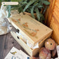 Large sized wooden pine personalised keepsake box. The design theme is perfect for New babies, Welcome to the world, baby showers and christenings. This design has baby bunnies, playing in a meadow style grass and a buzzy Bee flying around. The picture also shows an example in a nursey room filled with nappies, vests and teddys. 