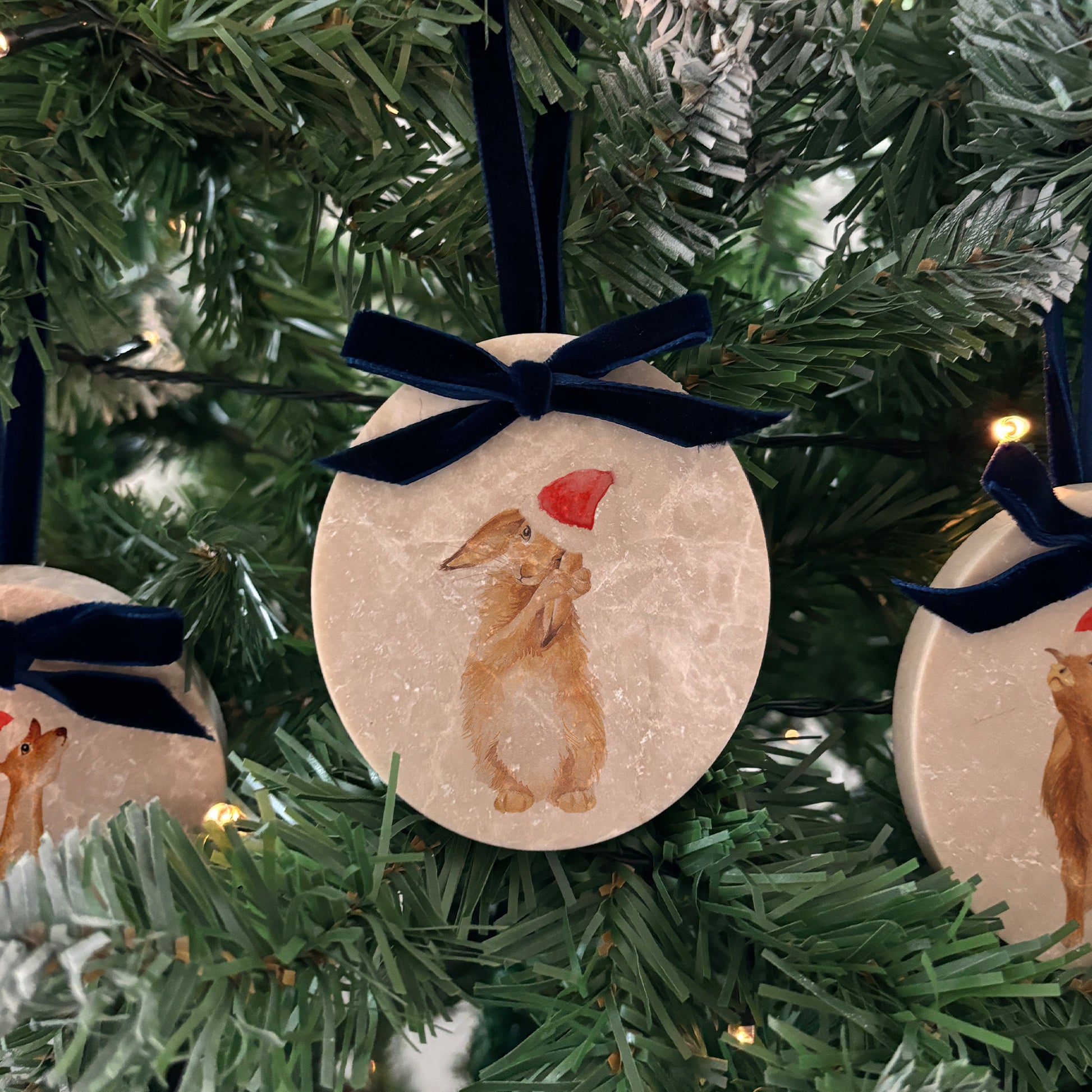 An oval marble bauble featuring a watercolour baby Humble Hare, with the option to add personalised text to celebrate baby's first Christmas, and to add a Santa hat. The bauble is finished with a velvet ribbon.