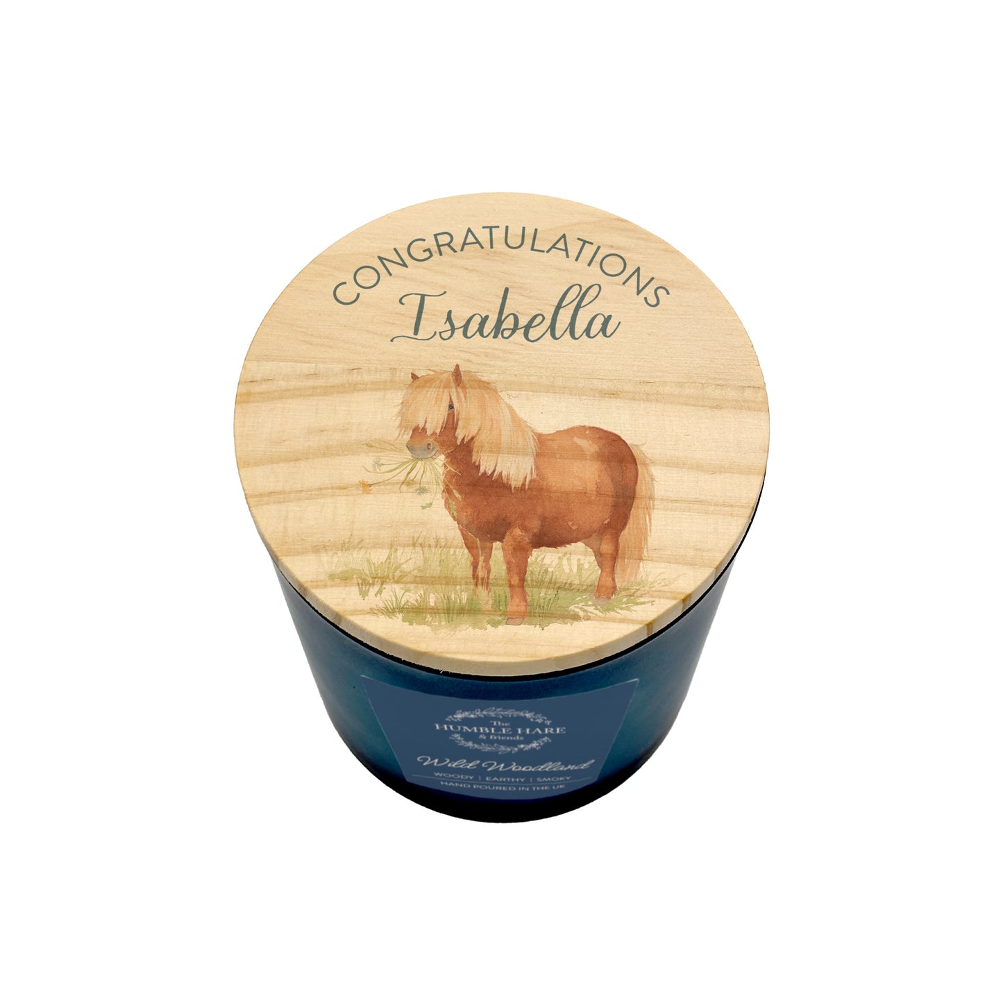 A personalised candle in a dark blue glass jar with a round wooden lid. The lid has 'CONGRATULATIONS Isabella' and features an illustration of a brown Shetland pony holding flowers in its mouth and standing in a grassy field.