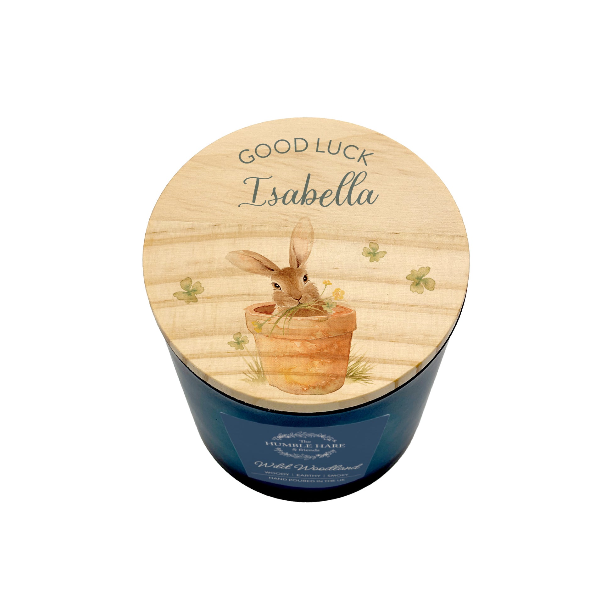 A personalised blue candle in a dark blue glass jar with a light wood lid with a bunny in a pot, clovers, and the words 'GOOD LUCK Isabella'.