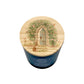 A blue cylindrical candle glass jar with a round wooden lid decorated with an illustration of a small rabbit sitting in front of a floral archway and arched door.